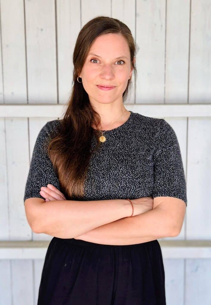 Christiane steht vor einer hellen Holzwand und hat die Arme verschränkt. Sie hat braune lange Haare und grüne Augen.