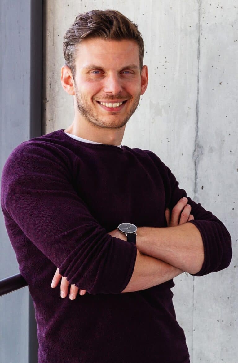 Tobias Roppelt steht vor einer grauen Wand und lacht in die Kamera. Er hat einen roten Pullover, braune Haare und blaue Augen.