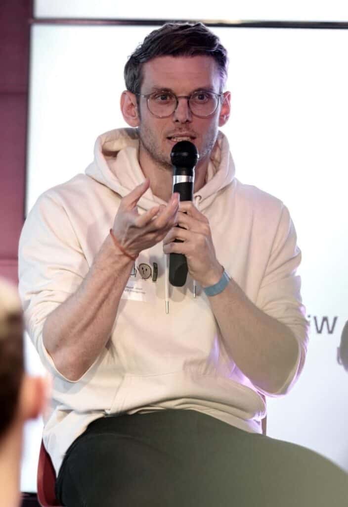 Tobias Roppelt sitzt auf einem Stuhl mit einem Mikrofon in der Hand und hält eine Rede.